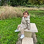 child, toddler, raincoat, outdoor, park, grass, stone_table, bench, greenery, trees, nature, casual_clothing, white_shoes, curly_hair, smile, sitting, young_child, daylight, casual_pose, playful
