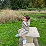 toddler, child, raincoat, white_shoes, picnic_table, stone_table, outdoor, grass, trees, nature, park, greenery, casual_clothing, curious, sitting, daytime, young_child, person, footwear, playful