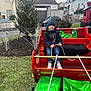 child, winter_clothing, hat, sleigh, presents, green_wrapping, red_sleigh, outdoor, grass, trees, houses, rope, smiling, jeans, shoes, cloudy_sky, holiday, festive, daytime, person