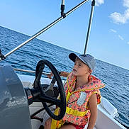 Swann participe au concours pour gagner de l'argent avec cette photo : child, life_jacket, boat, steering_wheel, ocean, water, sky, clouds, cap, summer, outdoor, nautical, vacation, marine, person, sitting, safety, recreation, transportation, youth
