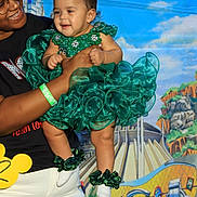 Loriana is registered to the contest to win money with this photo: adult, baby, background_mural, black_shirt, cartoon_characters, castle, celebration, child, costume, festive, glasses, green_dress, happy, headpiece, holding, outdoor, person, portrait, smile, white_pants