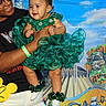 adult, baby, background_mural, black_shirt, cartoon_characters, castle, celebration, child, costume, festive, glasses, green_dress, happy, headpiece, holding, outdoor, person, portrait, smile, white_pants