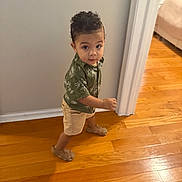 Theon is registered to the contest to win money with this photo: toddler, child, curly_hair, green_shirt, shorts, sandals, wooden_floor, indoor, walking, looking_up, doorframe, wall, home, young_child, person, casual_clothing, small_footwear, cute, curious, standing