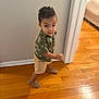 toddler, child, curly_hair, green_shirt, shorts, sandals, wooden_floor, indoor, walking, looking_up, doorframe, wall, home, young_child, person, casual_clothing, small_footwear, cute, curious, standing