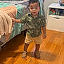 Theon joined the competition — help win amazing prizes! toddler, child, standing, bed, blanket, quilt, bedroom, floor, wooden_floor, furniture, nightstand, clothing, shirt, shorts, sandals, person, indoor, curious, holding, home