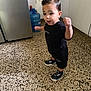 toddler, child, black_clothing, sneakers, curly_hair, indoor, kitchen, water_dispenser, floor_tiles, cabinet, expression, motion, young_child, person, standing, black_shorts, black_tshirt, playful, happy