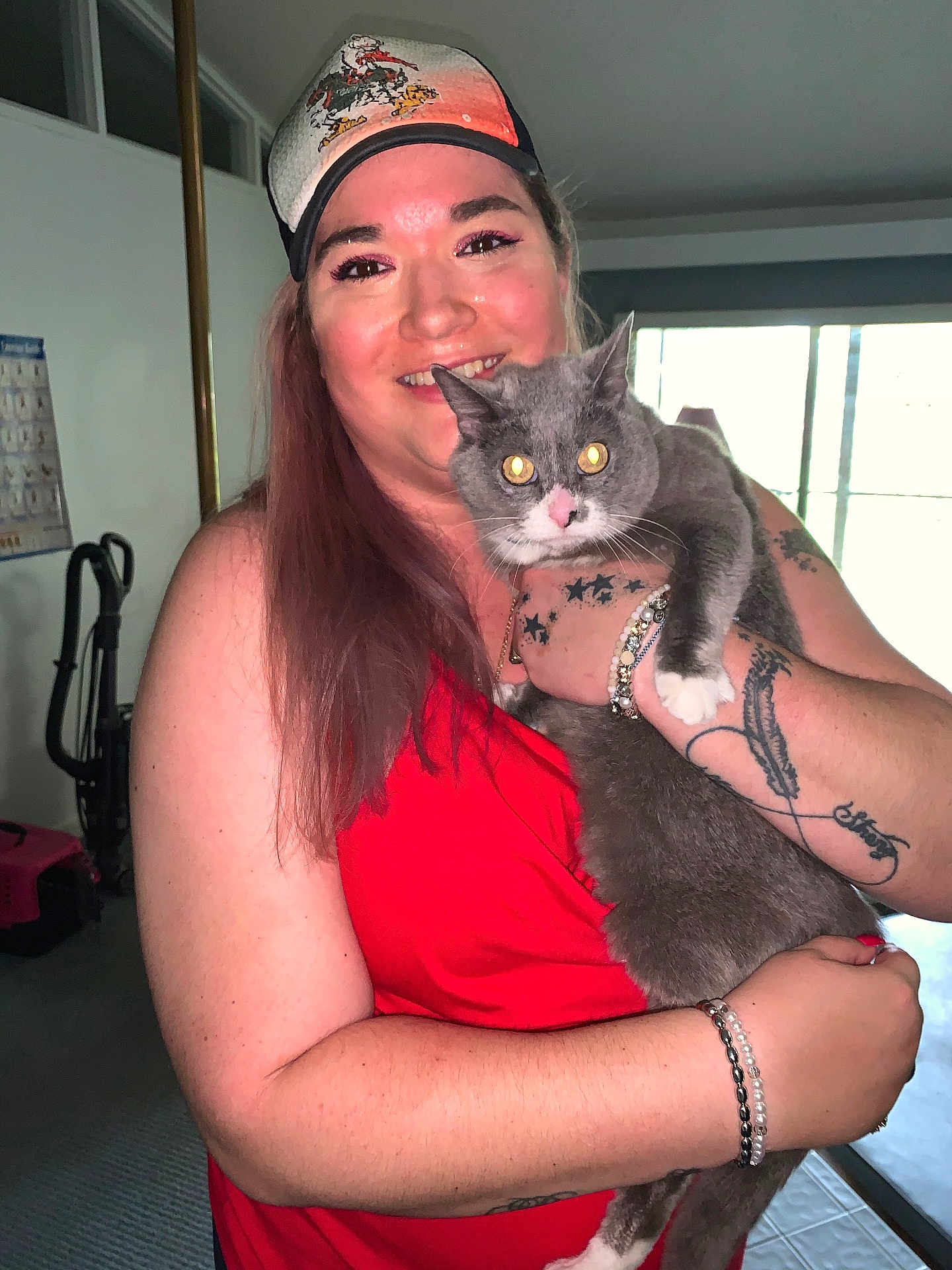 Martini Coons is registered to the contest to win money with this photo: bracelet, cap, cat, face, gray_cat, happy, holding, home_interior, indoor, long_hair, natural_light, person, pet, portrait, red_top, smiling, tattoo, vacuum_cleaner, white_paws, window
