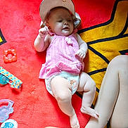 Riley is registered to the contest to win money with this photo: baby, infant, pink_top, diaper, hat, red_blanket, yellow_pattern, toy, pacifier, container, leg, child, person, smiling, lying_down, indoors, colorful, playful, cute, relaxed