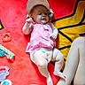 baby, infant, pink_top, diaper, hat, red_blanket, yellow_pattern, toy, pacifier, container, leg, child, person, smiling, lying_down, indoors, colorful, playful, cute, relaxed