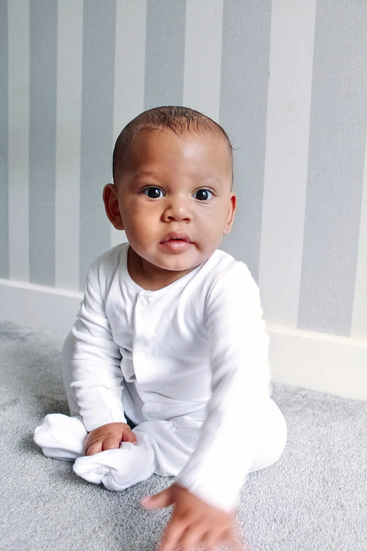 Kyrie is registered to the contest to win money with this photo: baby, child, infant, pajamas, white_clothing, carpet, floor, indoor, portrait, curious, reaching_hand, striped_wallpaper, home, cute, young_child, sitting, soft_lighting, person, expression, cozy