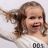 Ambre a rejoint le concours — aidez-le/la à gagner de superbes lots ! child, girl, curly_hair, smile, white_top, number_tag, portrait, happy, playful, light_background, one_shoulder, cute, face, young, blue_eyes, expression, hand, studio, fashion, model