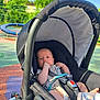 baby, canopy, casual_clothing, child, colorful_ground, daylight, footwear_free, greenery, infant, nature, outdoor, park, plastic_bottle, playground, relaxing, safety_harness, seatbelt, stroller, summer, sunlight