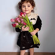 Larissa participe au concours pour gagner de l'argent avec cette photo : child, girl, tulips, flowers, bouquet, curly_hair, black_dress, lace_collar, smile, shoes, standing, indoor, portrait, cute, young, flower_stems, floor, wall, person, happy