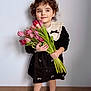 child, girl, tulips, flowers, bouquet, curly_hair, black_dress, lace_collar, smile, shoes, standing, indoor, portrait, cute, young, flower_stems, floor, wall, person, happy