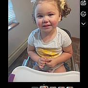 Nya Robbie is registered to the contest to win money with this photo: toddler, child, smiling, curly_hair, high_chair, indoor, window, white_tshirt, yellow_sun, hair_bows, hands, face, person, young_child, happy, seated, floor_tiles, natural_light, home, casual_clothing