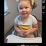 toddler, child, smiling, curly_hair, high_chair, indoor, window, white_tshirt, yellow_sun, hair_bows, hands, face, person, young_child, happy, seated, floor_tiles, natural_light, home, casual_clothing