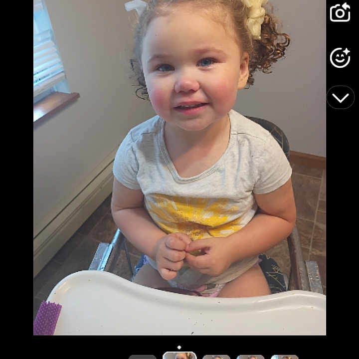 Nya Robbie is registered to the contest to win money with this photo: casual_clothing, child, curly_hair, face, floor_tiles, hair_bows, hands, happy, high_chair, home, indoor, natural_light, person, seated, smiling, toddler, white_tshirt, window, yellow_sun, young_child