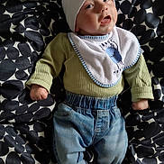 Ellis is registered to the contest to win money with this photo: baby, infant, child, hat, bib, green_sweater, blue_jeans, lying_down, blanket, patterned_blanket, clothing, cute, face, portrait, indoors, small_hands, footwear_absent, expression, looking_up, casual