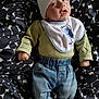 baby, infant, child, hat, bib, green_sweater, blue_jeans, lying_down, blanket, patterned_blanket, clothing, cute, face, portrait, indoors, small_hands, footwear_absent, expression, looking_up, casual