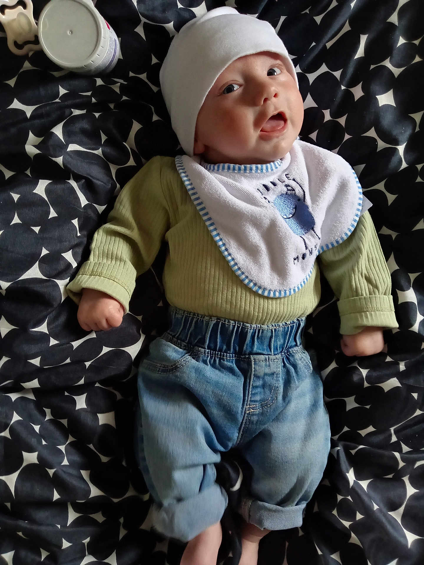Ellis is registered to the contest to win money with this photo: baby, infant, child, hat, bib, green_sweater, blue_jeans, lying_down, blanket, patterned_blanket, clothing, cute, face, portrait, indoors, small_hands, footwear_absent, expression, looking_up, casual