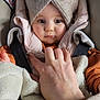 baby, child, headwrap, blanket, car_seat, hand, clothing, face, person, warm, seatbelt, orange_sweater, soft_texture, indoors, closeup, infant, cute, expression, protective, bundle