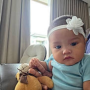 Carissa is registered to the contest to win money with this photo: baby, child, plush_toy, teddy_bear, headband, flower, indoor, couch, curtains, window, hand, person, clothing, blue_shirt, face, expressionless, furniture, soft_toy, seated, natural_light