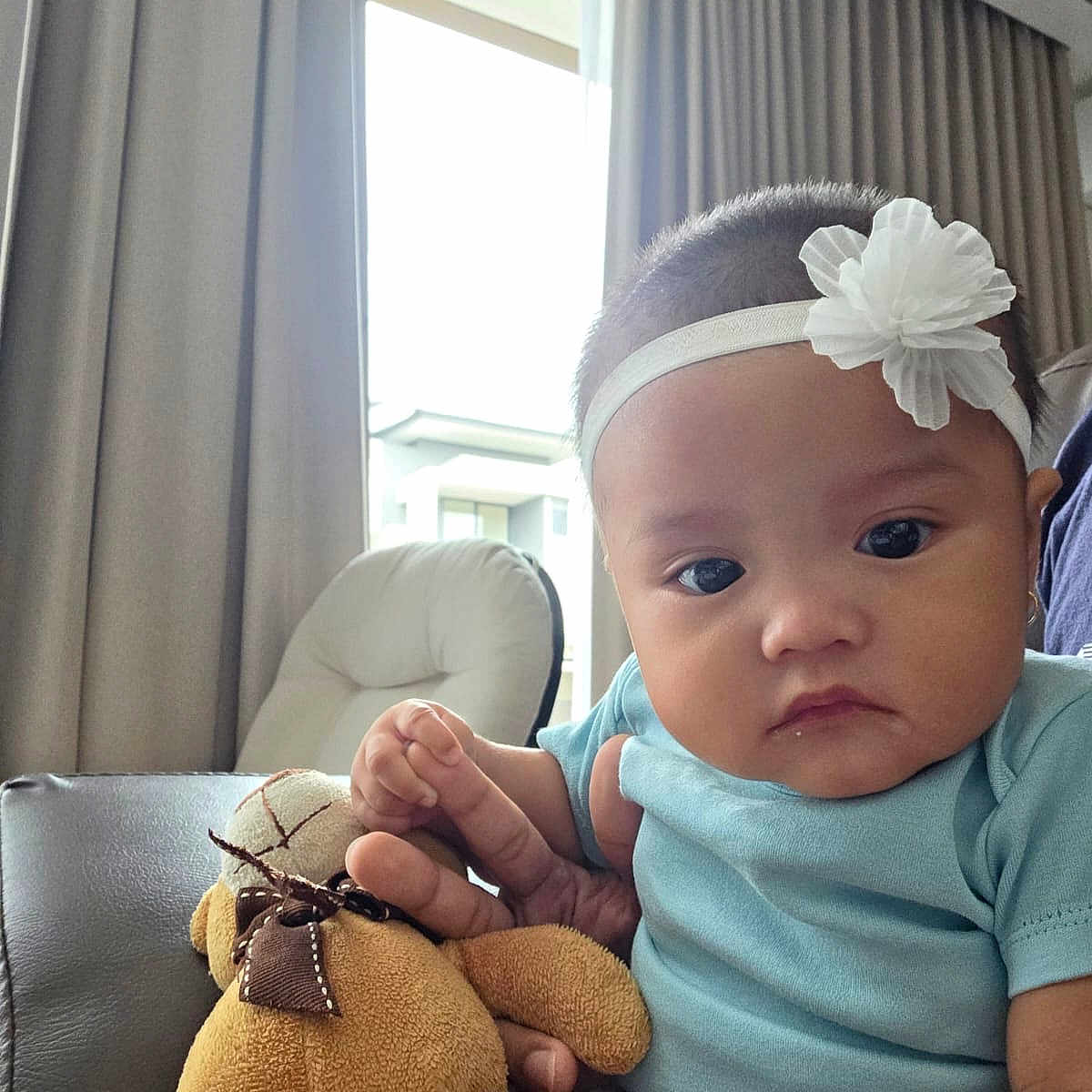 Carissa is registered to the contest to win money with this photo: baby, blue_shirt, child, clothing, couch, curtains, expressionless, face, flower, furniture, hand, headband, indoor, natural_light, person, plush_toy, seated, soft_toy, teddy_bear, window