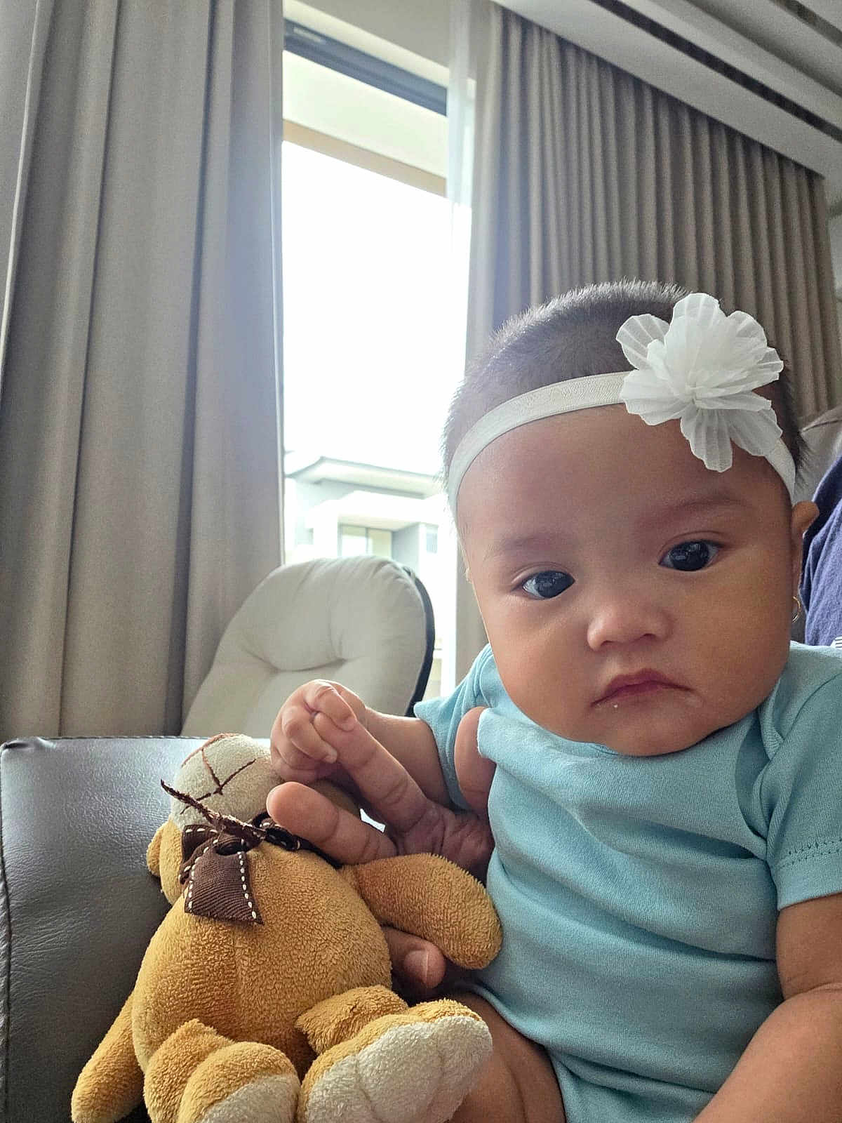 Carissa is registered to the contest to win money with this photo: baby, child, plush_toy, teddy_bear, headband, flower, indoor, couch, curtains, window, hand, person, clothing, blue_shirt, face, expressionless, furniture, soft_toy, seated, natural_light