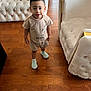 child, toddler, standing, indoor, living_room, sofa, wooden_floor, curly_hair, shorts, tshirt, sneakers, furniture, cushion, expression, person, casual_clothing, home_interior, smiling, cute, looking_up