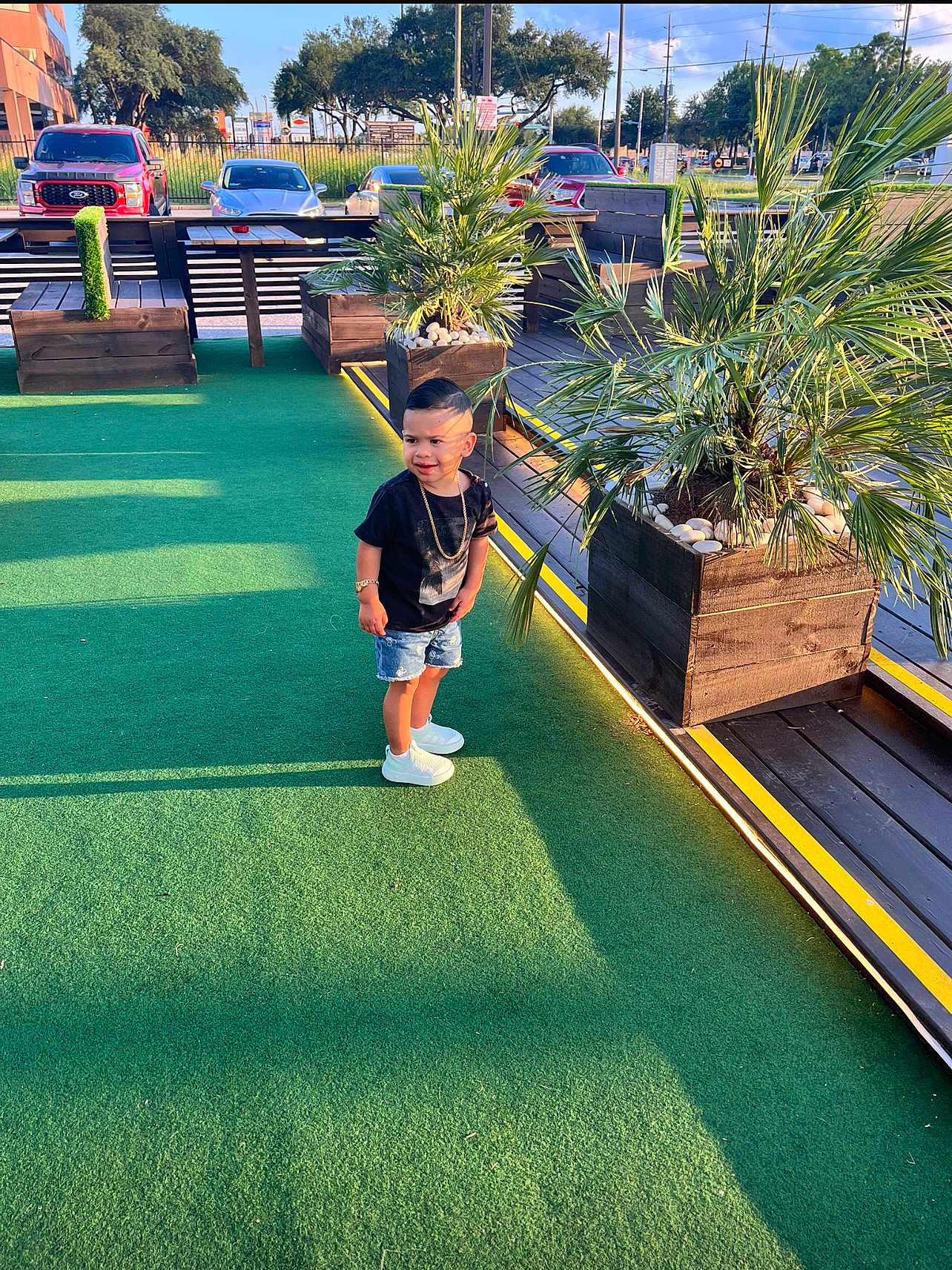 Aron is registered to the contest to win money with this photo: toddler, child, outdoor, green_carpet, plant, palm, wooden_planter, sunlight, shadow, casual_clothing, denim_shorts, white_sneakers, black_tshirt, gold_chain, bracelet, parking_lot, car, sky, daytime, sidewalk
