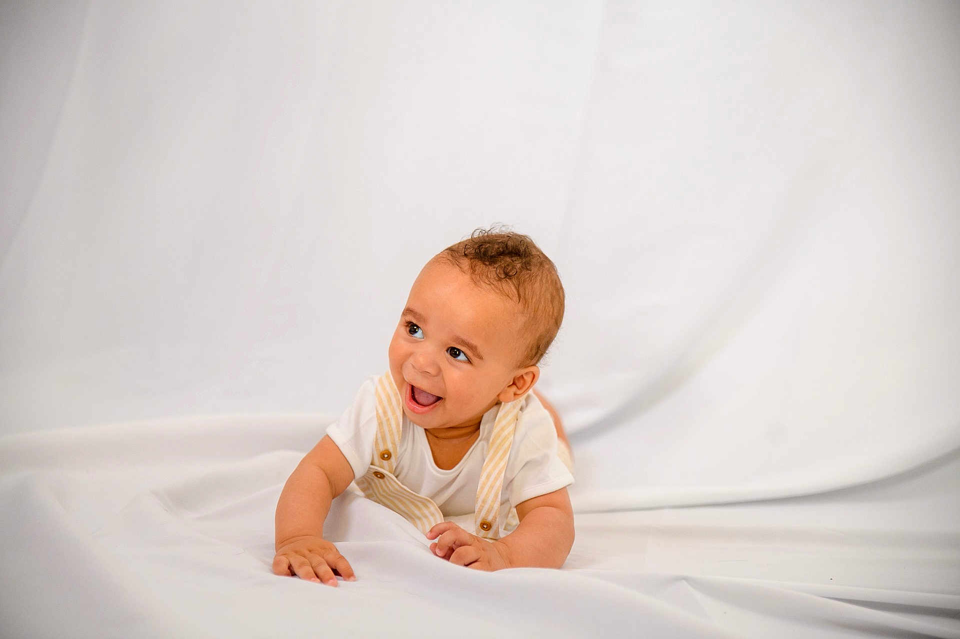 Ruben joined the competition — help win amazing prizes! baby, infant, child, smiling, happy, lying_down, white_background, overalls, curly_hair, cute, portrait, one_person, indoors, soft_lighting, toddler, skin, face, hands, white_clothing, expression