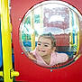 child, playground, bubble_window, smile, happy, plastic, red_panel, yellow_pole, outdoor, play, fun, young_child, face, hand, casual_clothing, daylight, recreation, park, curious, cute