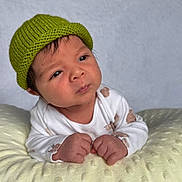 Salim joined the competition — help win amazing prizes! baby, infant, hat, knitted_hat, green_hat, onesie, white_onesie, brown_spots, cushion, soft_texture, yellow_cushion, face, hands, curious, indoors, portrait, child, newborn, skin, expression