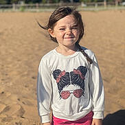 Kalia is registered to the contest to win money with this photo: braid, casual_clothing, child, daylight, fence, girl, graphic_tee, nature, outdoor, pink_pants, playground, portrait, sand, smile, squinting, standing, sunlight, trees, white_sweatshirt, young