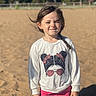 braid, casual_clothing, child, daylight, fence, girl, graphic_tee, nature, outdoor, pink_pants, playground, portrait, sand, smile, squinting, standing, sunlight, trees, white_sweatshirt, young