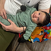 Mason is registered to the contest to win money with this photo: adult_arm, baby, black_mittens, brown_leather_couch, child, colorful_blanket, couch, cute, dinosaur_outfit, green_clothing, happy, holding, indoor, infant, m_and_m_pattern, person, portrait, smile, toddler, white_tshirt
