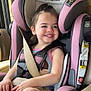 child, toddler, car_seat, seatbelt, vehicle_interior, smiling, happy, safety, pink, gray, clothing, hair, skin, face, seat, window, daylight, armrest, hands, legs
