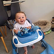 Alessio participe au concours pour gagner de l'argent avec cette photo : baby, walker, blue, white, toy, steering_wheel, smiling, happy, indoor, floor, tile, chair, pet_bed, foot, person, child, cute, hair, clothing, home
