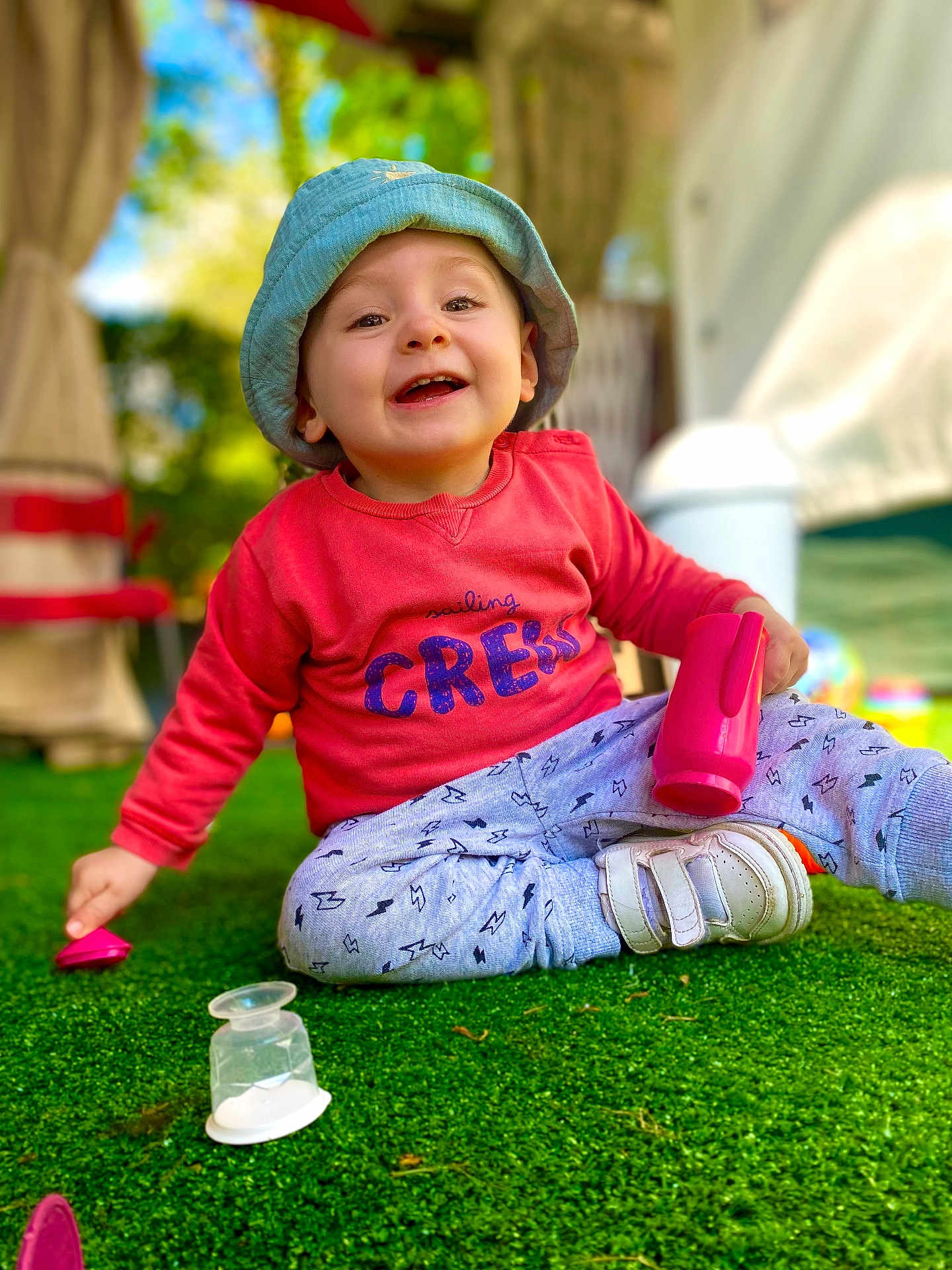 Kaylan participe au concours pour gagner de l'argent avec cette photo : toddler, child, smiling, hat, bucket_hat, sweatshirt, pink, toy, grass, outdoor, daylight, happy, playing, sitting, footwear, white_shoes, casual_clothing, blurred_background, portrait, closeup