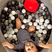 Kaylan a rejoint le concours — aidez-le/la à gagner de superbes lots ! baby, child, pacifier, ball_pit, balls, toy_ball, striped_clothing, arms_outstretched, indoor, flooring, grey_ball_pit, white_balls, black_balls, translucent_balls, red_ball, purple_ball, cute, lying_down, infant, playtime