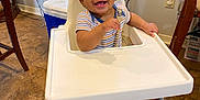 Davian is registered to the contest to win money with this photo: baby, high_chair, smiling, indoor, floor_tiles, plant, window, curtains, pacifier, slippers, baby_bear, child, person, happy, footrest, wooden, striped_shirt, home, furniture, cute