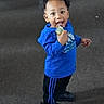 toddler, child, blue_hoodie, black_pants, standing, smiling, snack_pouch, indoor, carpet, happy, person, young_child, casual_clothing, footwear, curly_hair, cute, playful, bright_clothing, full_body, portrait