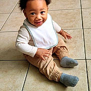 Panthir joined the competition — help win amazing prizes! toddler, child, smiling, sitting, curly_hair, bib, beige_pants, gray_socks, tile_floor, indoor, happy, cute, baby, person, portrait, young_child, casual_clothing, small_hands, bright_eyes, flooring