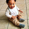 toddler, child, smiling, sitting, curly_hair, bib, beige_pants, gray_socks, tile_floor, indoor, happy, cute, baby, person, portrait, young_child, casual_clothing, small_hands, bright_eyes, flooring