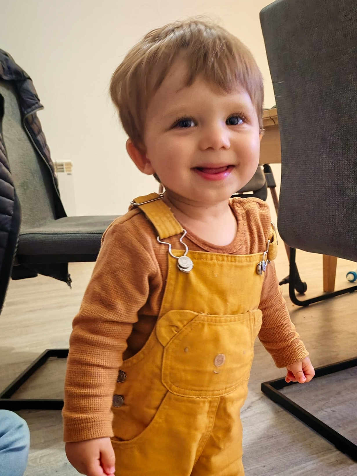 Raphael a rejoint le concours — aidez-le/la à gagner de superbes lots ! toddler, child, smiling, indoor, overalls, yellow_clothing, brown_clothing, face, happy, furniture, chair, table, floor, person, curly_hair, cute, standing, playful, young_child, expression