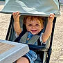 toddler, child, stroller, outdoor, happy, smiling, canopy, scraped_knee, denim_overalls, seatbelt, sunlight, playful, baby, person, casual_clothing, table, concrete, greenery, daylight, baby_stroller