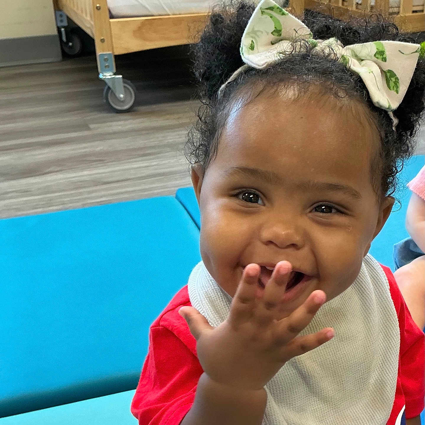 Leliana joined the competition — help win amazing prizes! toddler, child, smiling, curly_hair, bow, bib, red_shirt, blue_mat, indoor, crib, wooden_crib, flooring, happy, person, baby, playful, cute, portrait, infant, room