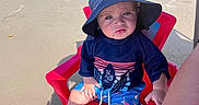 Karson joined the competition — help win amazing prizes! baby, child, beach, sand, sun_hat, red_chair, seated, pouting, portrait, outdoor, sunlight, coastline, dunes, boardwalk, ocean, summer, swimwear, blue_clothing, barefoot, cloudy_sky