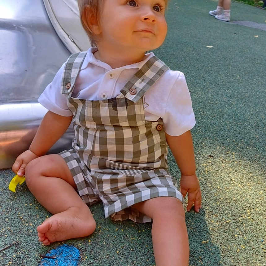 Itao a rejoint le concours — aidez-le/la à gagner de superbes lots ! asphalt, barefoot, child, curious, cute, daylight, greenery, outdoor, overalls, park, person, play_area, playground, sitting, slide, summer, toddler, trash_bin, white_shirt, young_child