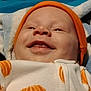 baby, blanket, child, closeup, cozy, cute, expression, face, happy, headwear, indoor, infant, lying_down, orange_hat, orange_pattern, portrait, smiling, soft_texture, warm, white_clothing